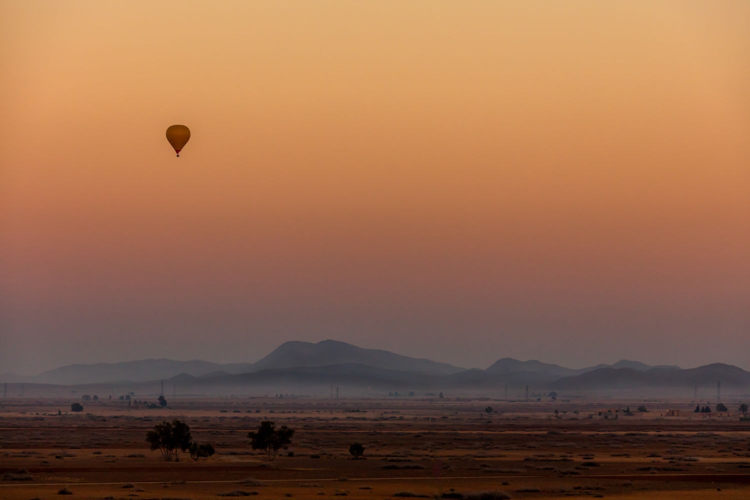 A hot air balloon silhouetted against a dusty orange sky during sunset over a desert plain with mountains in the distance.