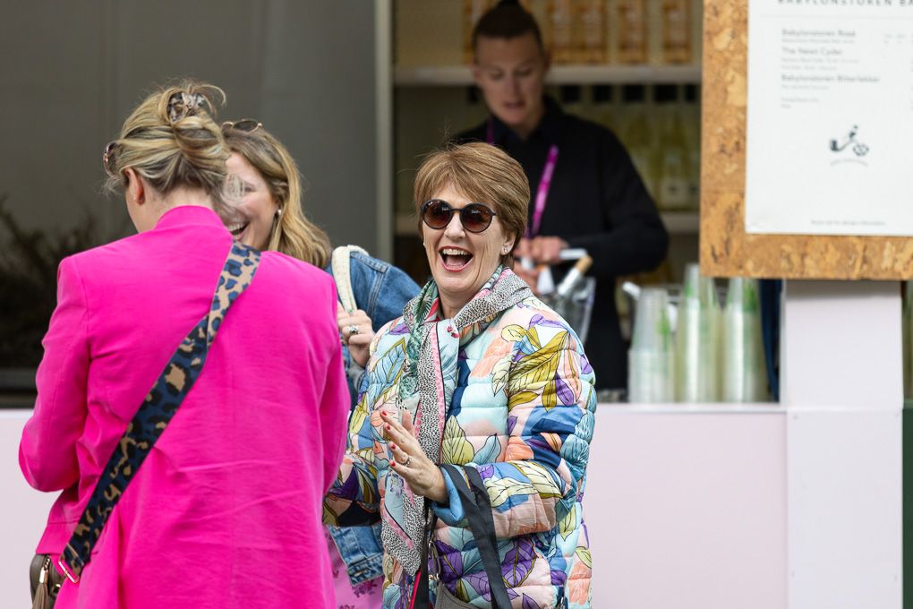 A person wearing a bright pink jacket and leopard print bag stands outside while others gather in the background.