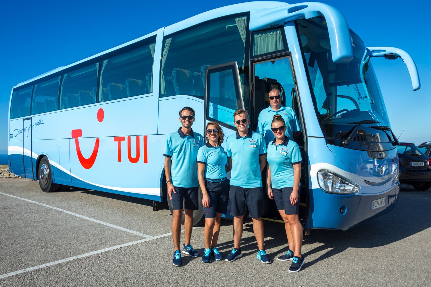 TUI staff wearing light blue uniforms pose in front of a TUI coach with a big red TUI Smile logo against a bright blue sky.