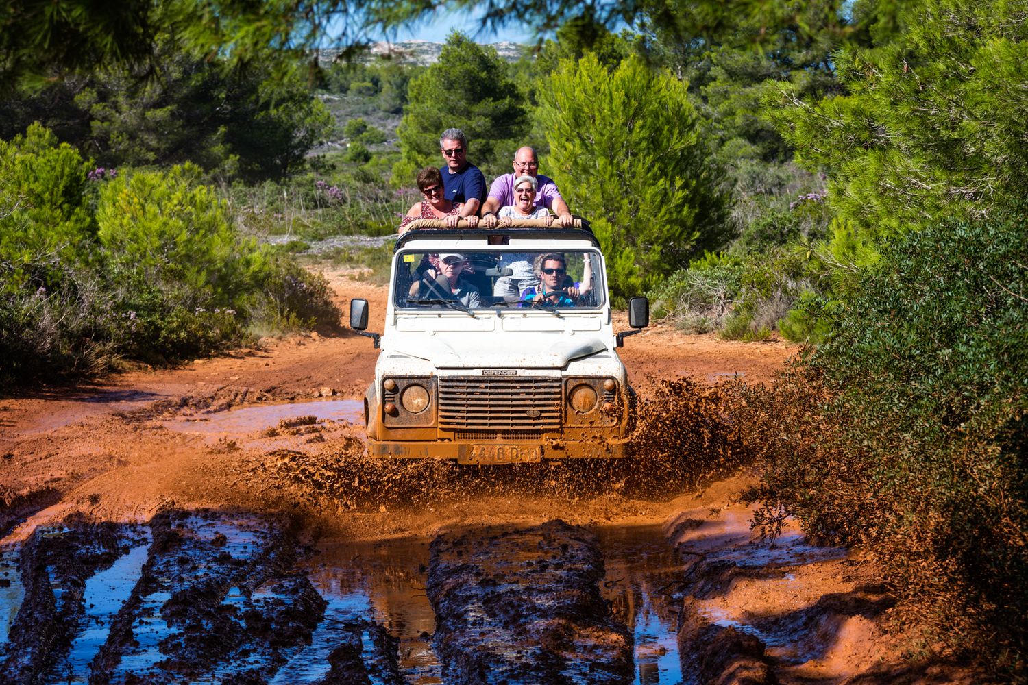 White off-road vehicle splashing through muddy water on a dirt trail surrounded by greenery.