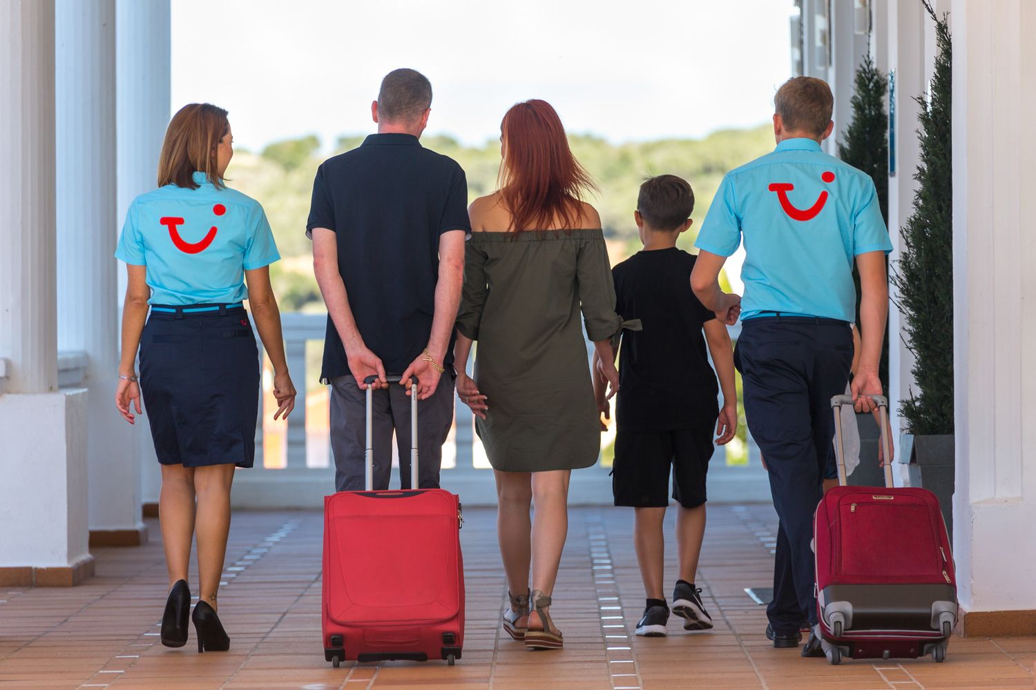 A group of travellers and TUI reps in uniform walking through a hotel corridor pulling rolling luggage suitcases.
