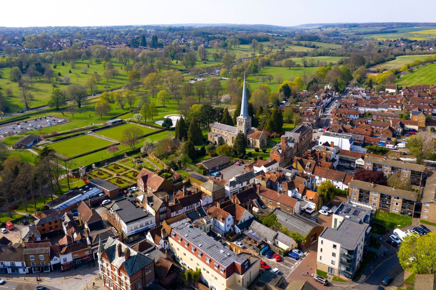 Aerial view of a charming English village with a church spire, red-roofed buildings, and lush green fields beyond.