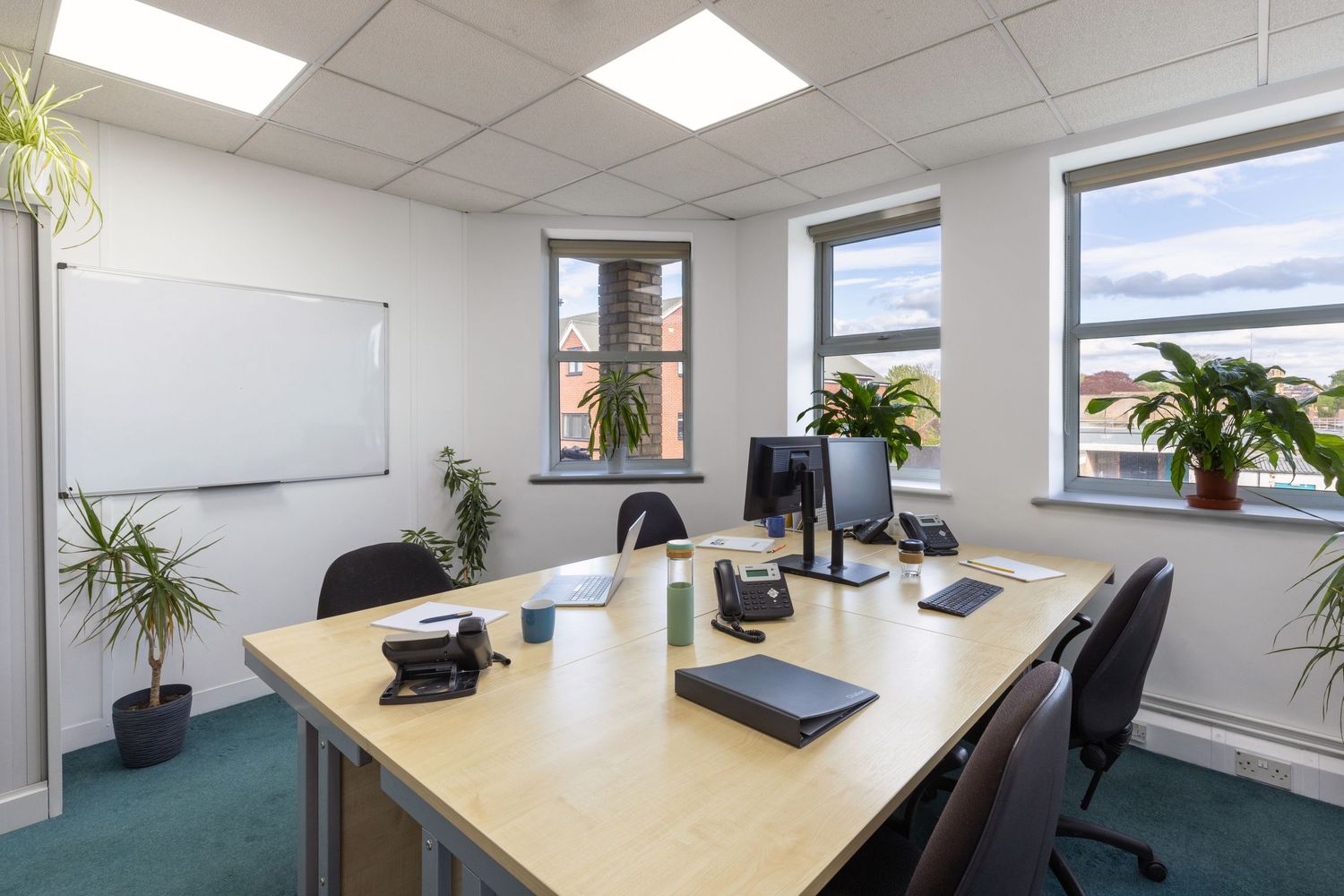 Modern office space with wooden desks, computer monitors, whiteboard, green plants, and natural light from large windows.