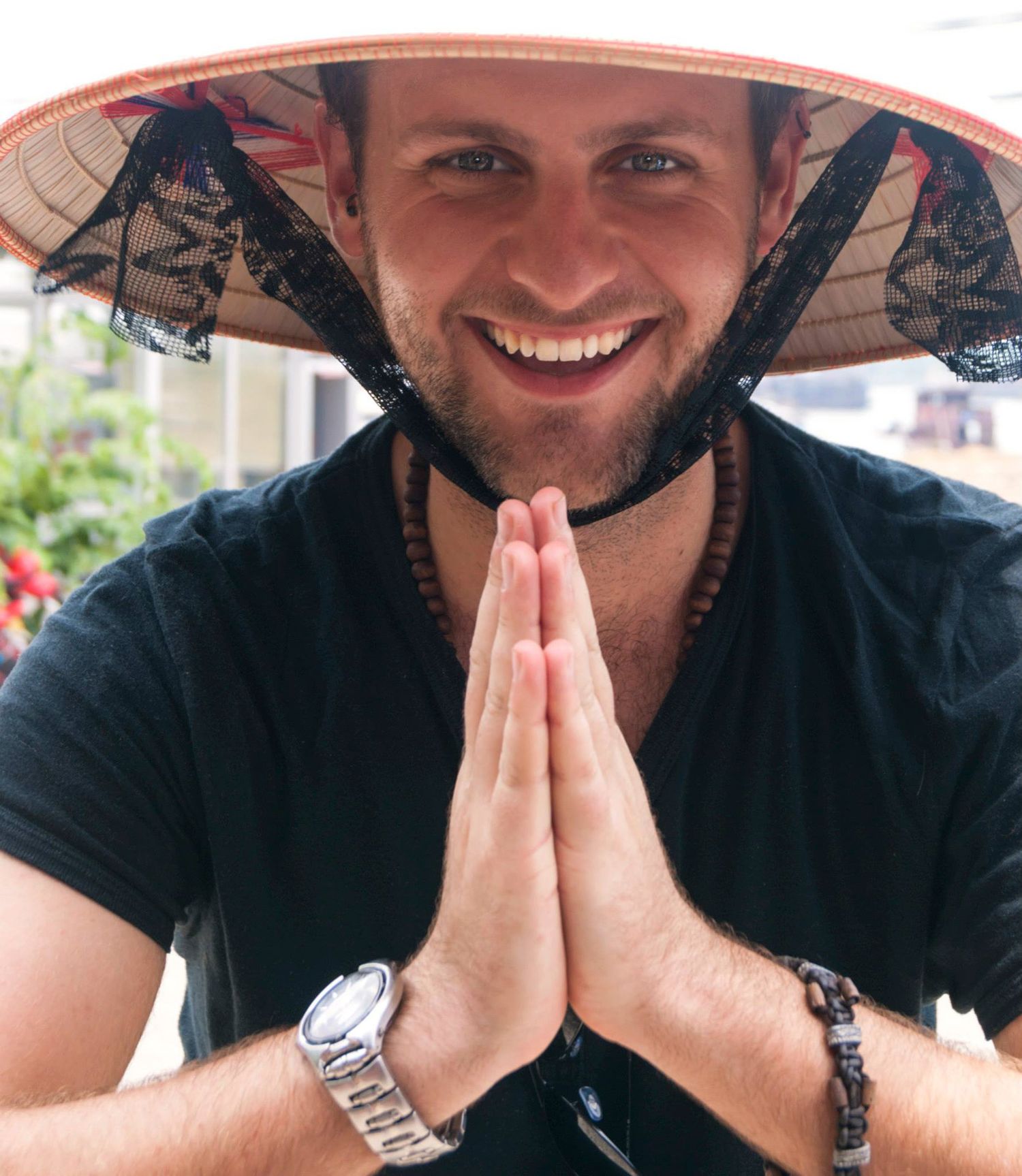A traveler wearing a conical Asian hat and black t-shirt holds their hands in prayer position while smiling.