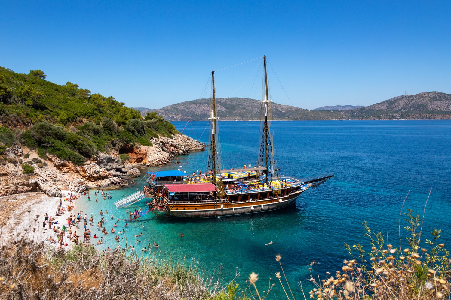 Traditional wooden sailing ship anchored in turquoise bay near rocky beach with mountains in background in Croatia.