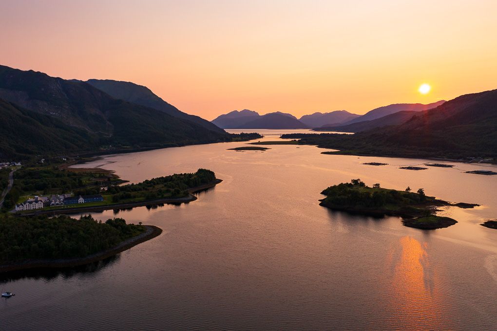 An aerial shot of a golden sunset over tranquil fjord waters with silhouetted mountains and islands in Glencoe, Scotland.