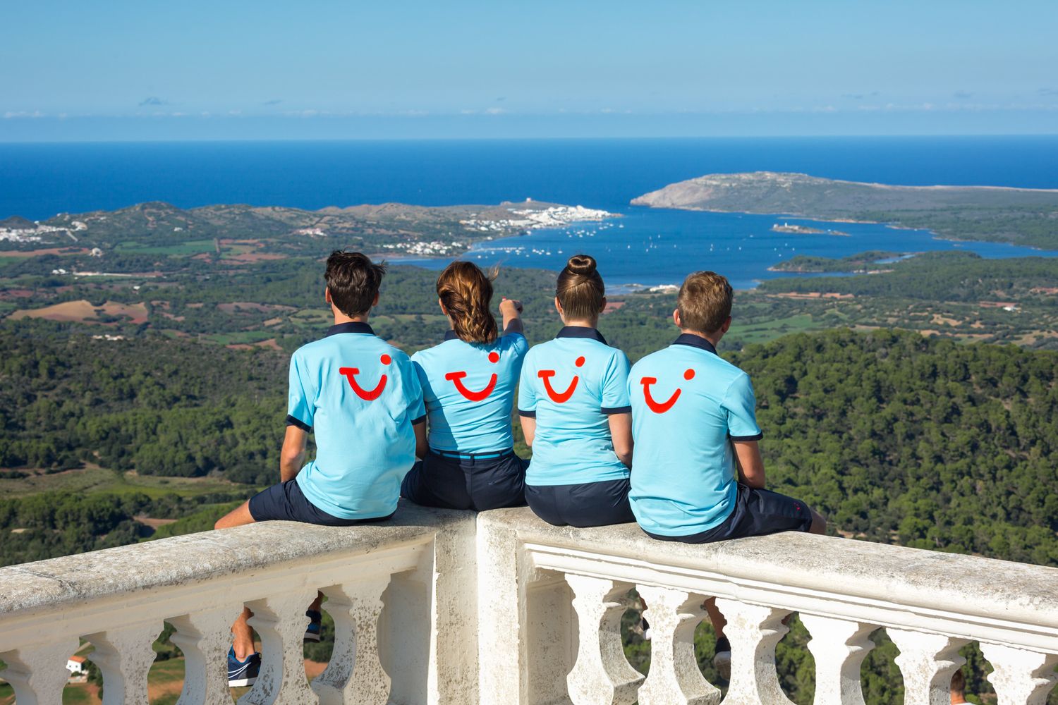 TUI reps in matching uniform with TUI Smile logo sit at a scenic mountain viewpoint overlooking the coastal Mediterranean landscape.