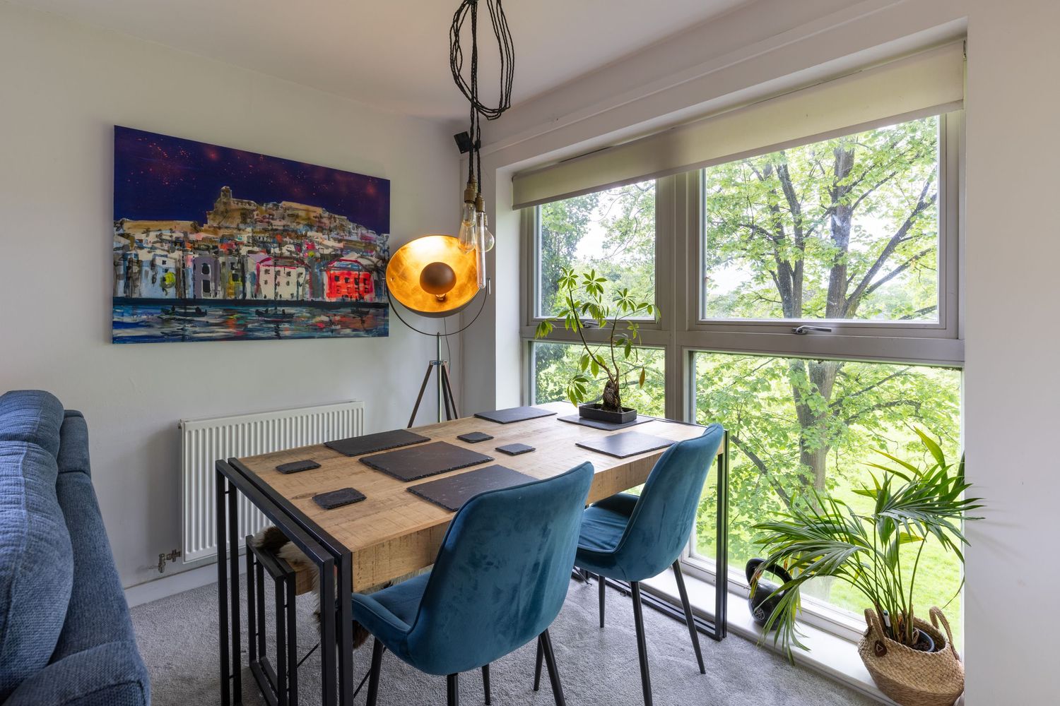 Modern dining area with blue velvet chairs, wooden table, colorful wall art, and large windows overlooking lush green trees.