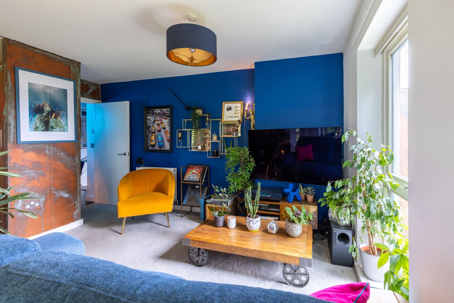 Vibrant living room with bold blue walls, yellow armchair, wooden coffee table, TV, and lush green plants by bright windows.