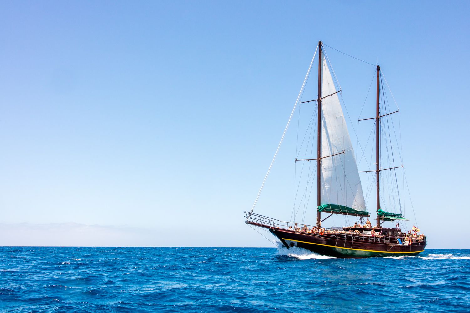 Traditional wooden sailboat with white sails cruising on bright blue tropical waters under clear skies.