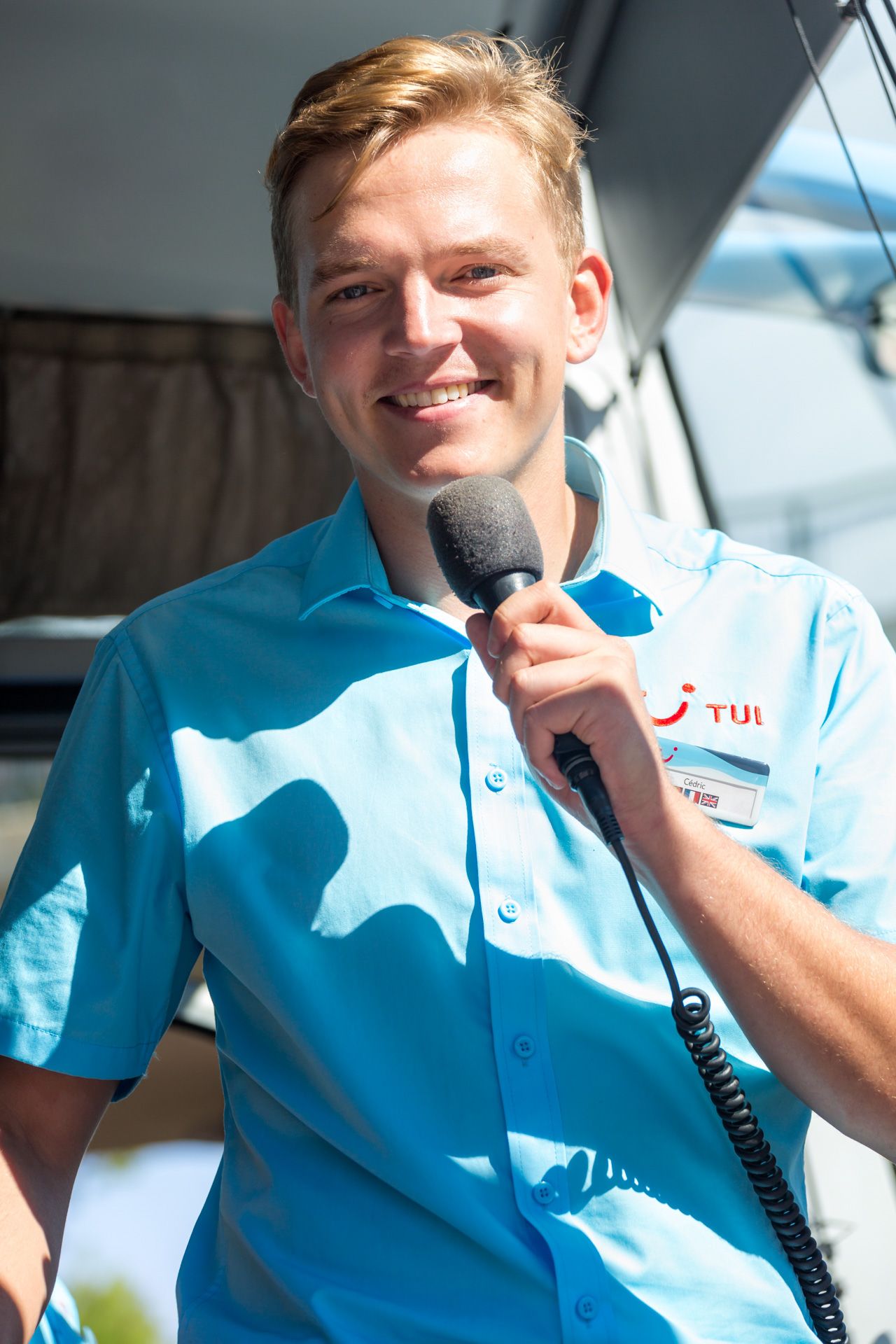 A tour representative in a light blue TUI uniform and red TUI Smile logo speaks into a microphone while smiling.