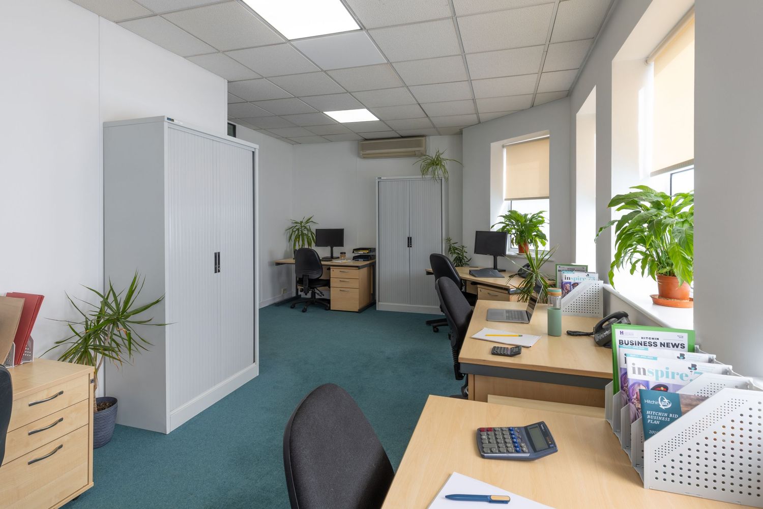 Modern office space with teal carpet, wooden desks, computers, green plants, and white storage cabinets under bright ceiling lights.