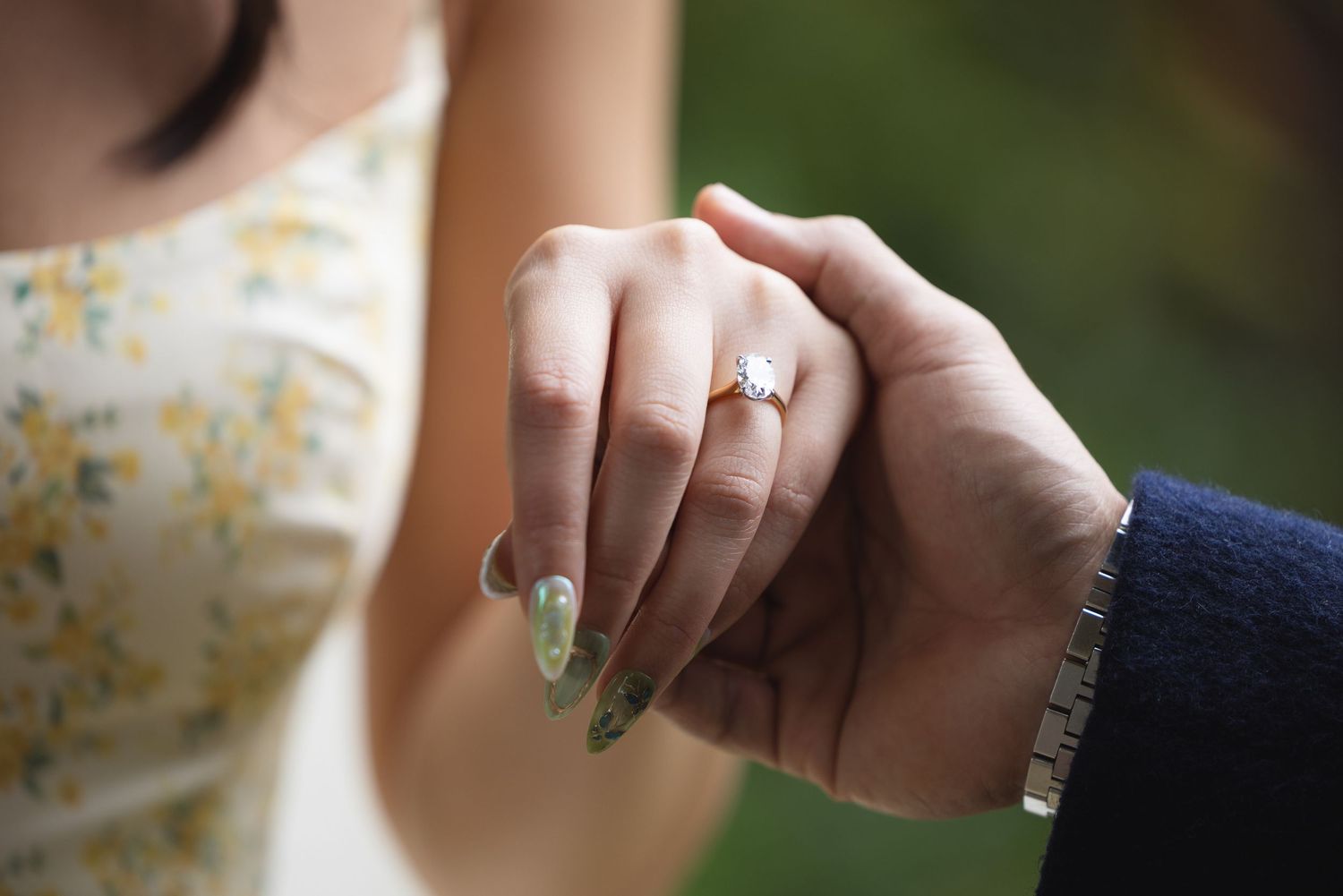 Close-up of a woman wearing a diamond engagement ring holding hands with a man in a navy suit.