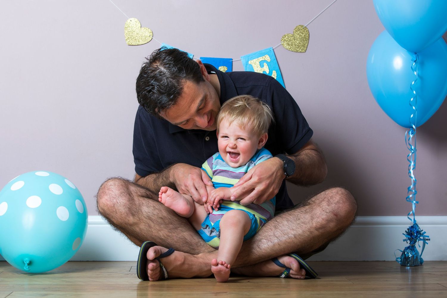 Father kissing laughing baby boy on first birthday with blue balloons and heart decorations in background.