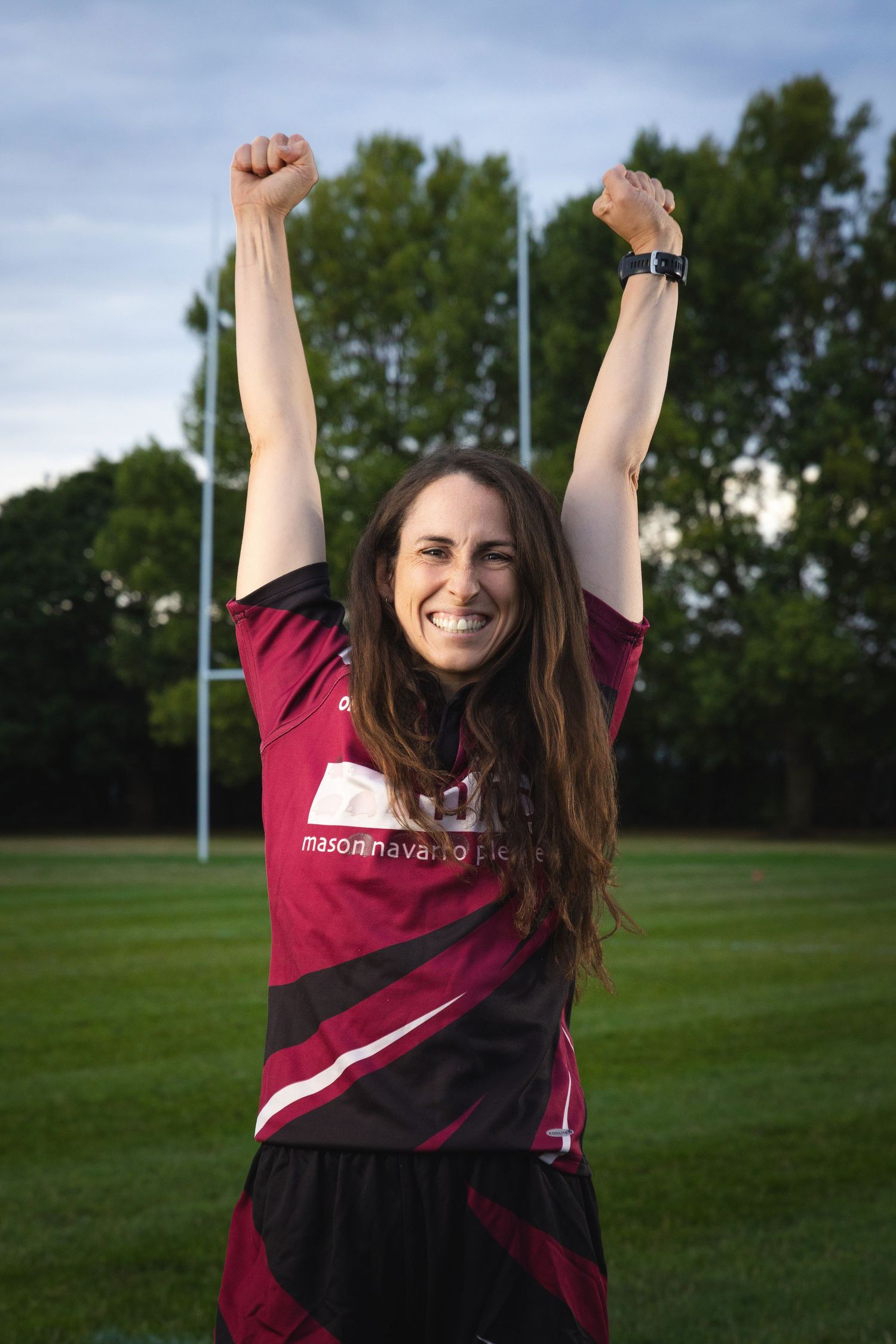 Woman in maroon sports jersey celebrates with arms raised on rugby field, smiling joyfully outdoors.