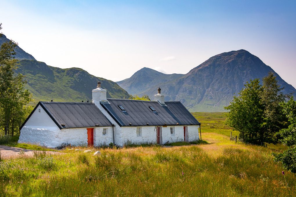 White farmhouse buildings nestled in a scenic mountain valley with golden grass fields and blue skies in Glencoe, Scotland.