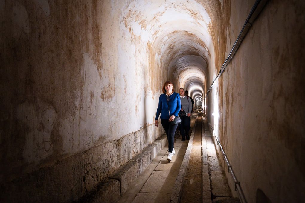 Narrow stone tunnel walkway with old weathered walls and dim lighting creates an atmospheric underground passageway.