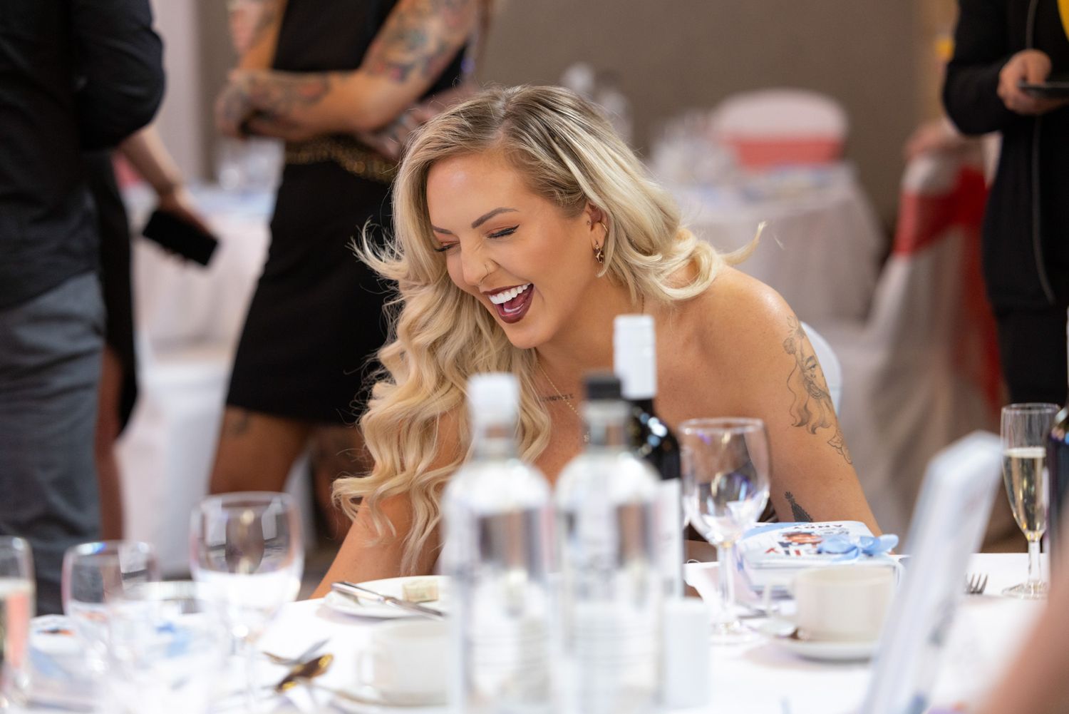 Event photography. A person in formal attire laughs joyfully while seated at an elegant dining table with glassware and place settings.