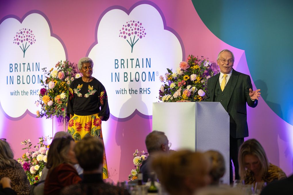 Event speakers stand at podium with Britain in Bloom signage and floral arrangements on stage during awards ceremony.