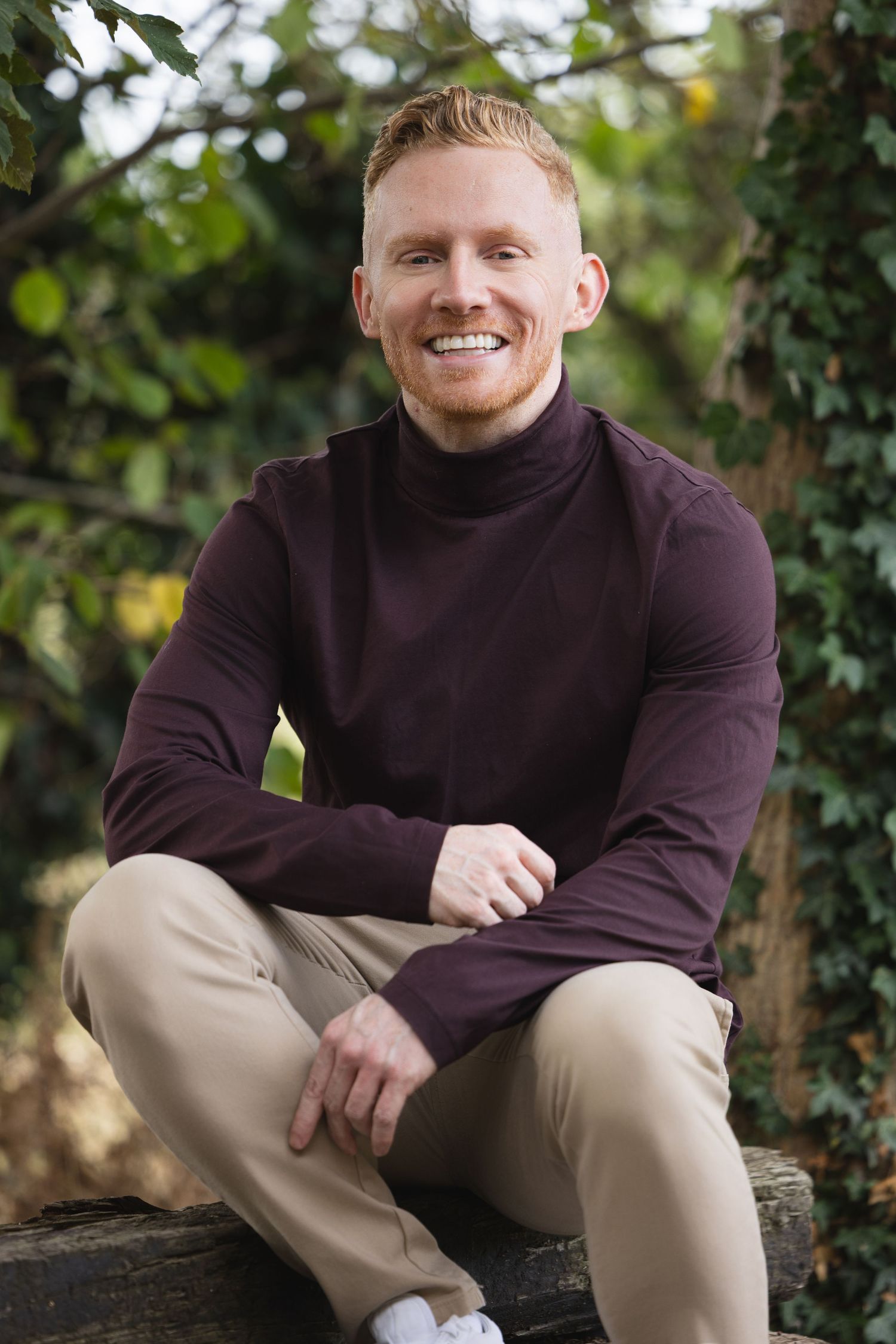 Smiling redhead man in maroon turtleneck and khaki pants sitting cross-legged on wooden bench in lush garden.