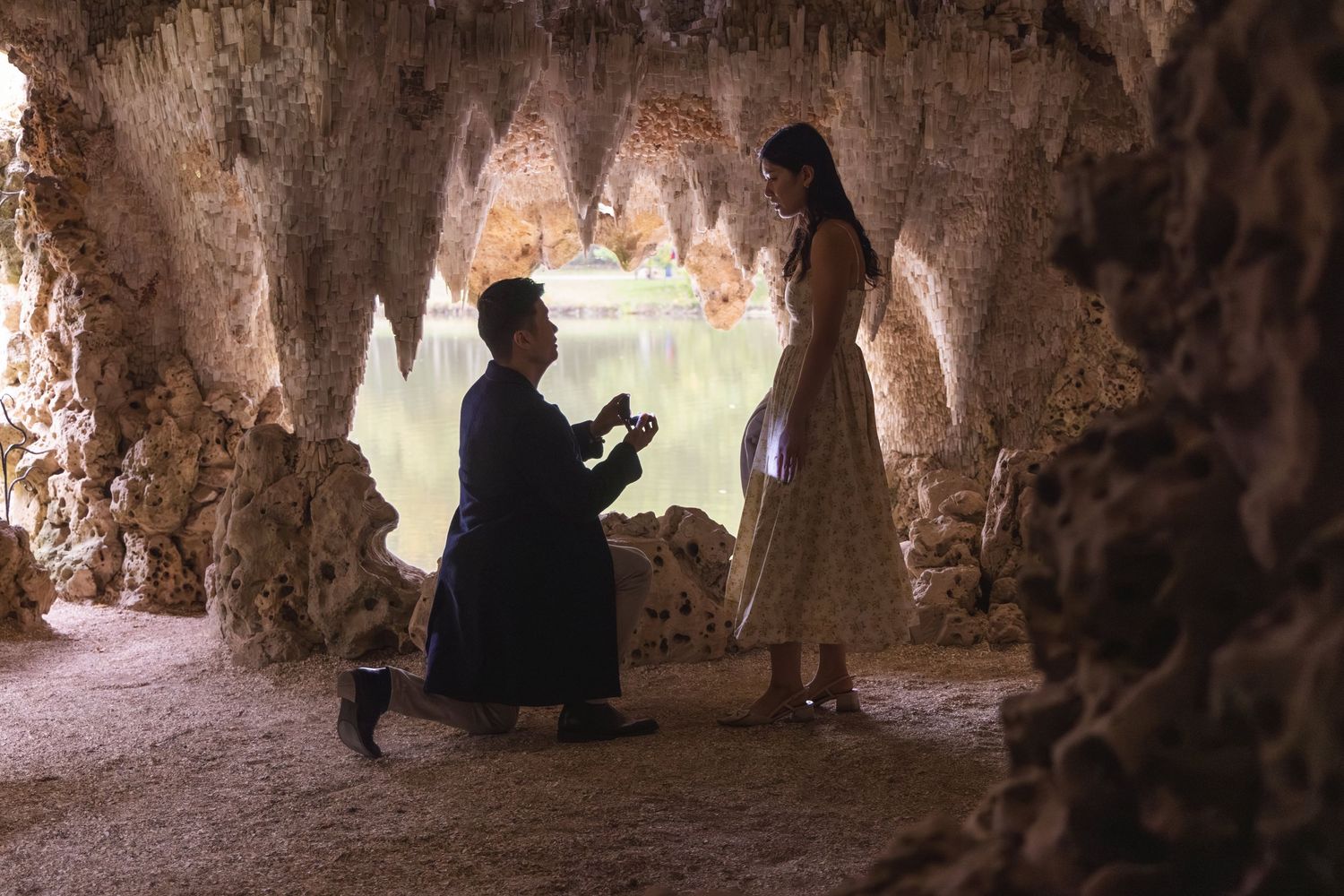 Man proposes to woman on one knee inside a stunning cave grotto with stalactites and a serene lake backdrop.