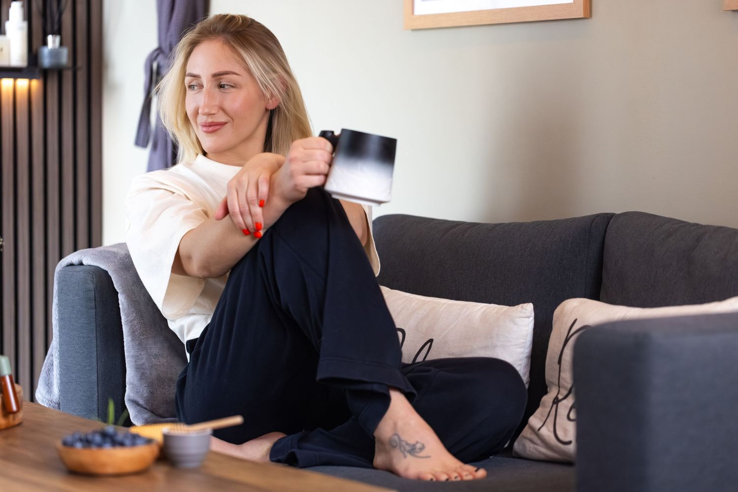 Woman relaxing on gray sofa holding black mug, wearing casual black pants, with tattoo on foot, near wooden coffee table.