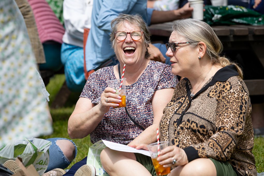 Press photography of two people enjoying drinks and laughing together outdoors at RHS Chelsea Flower Show in London