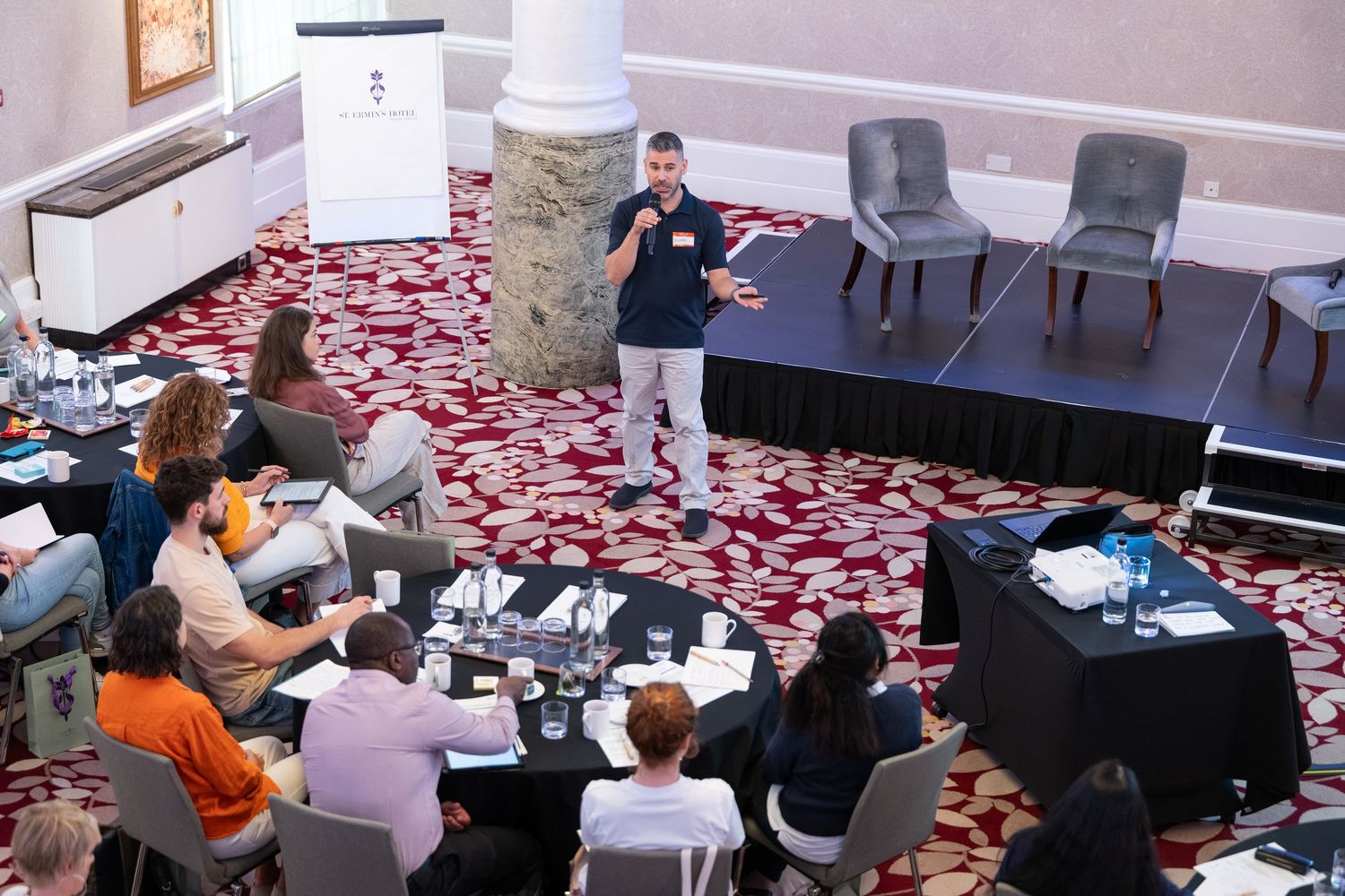 A speaker presents to a diverse audience seated at tables in an elegant hotel conference room with red patterned carpet.