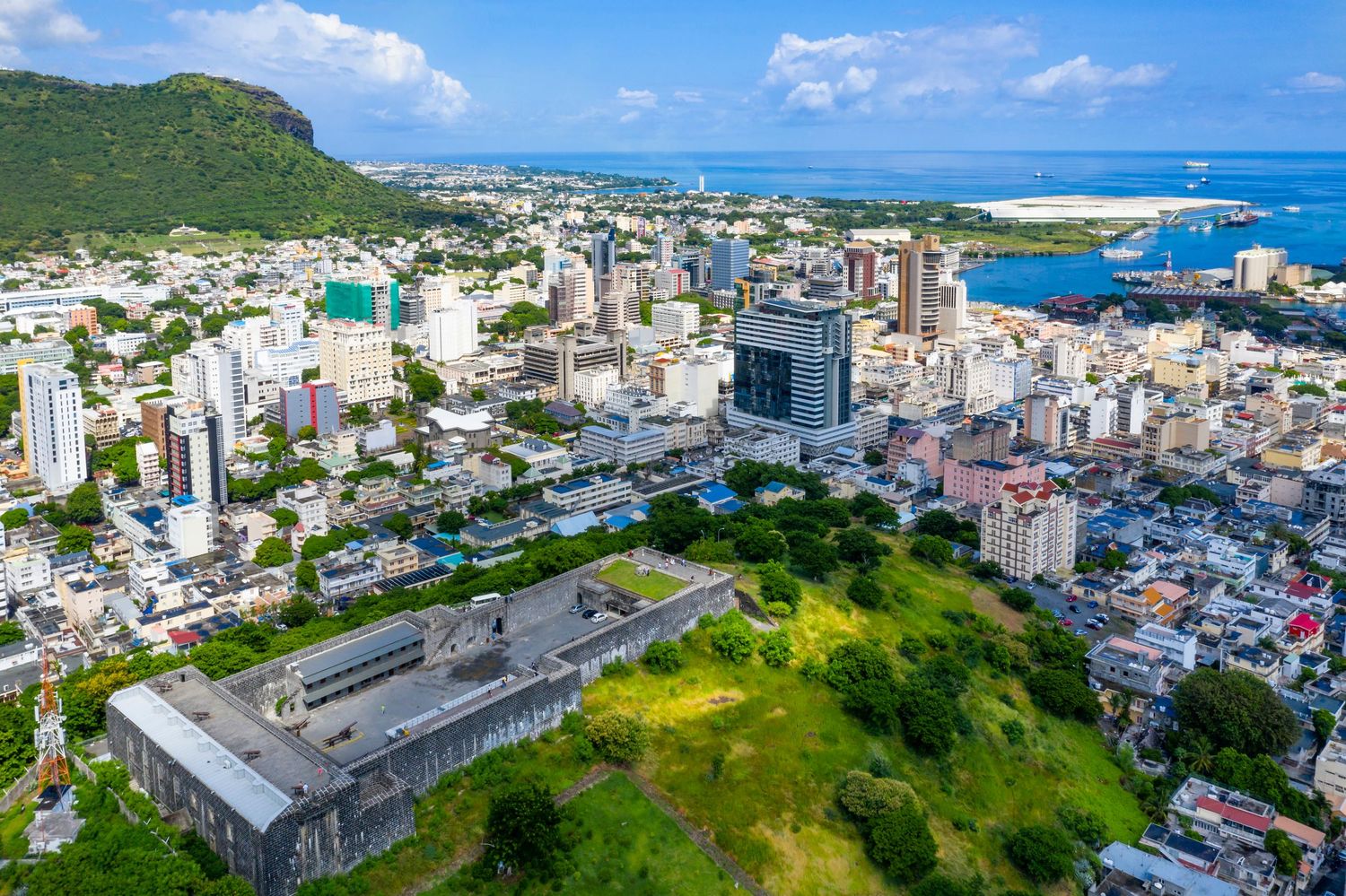 Aerial view of Port Louis, Mauritius, showcasing urban skyline, lush hills, and harbor under a clear blue sky.
