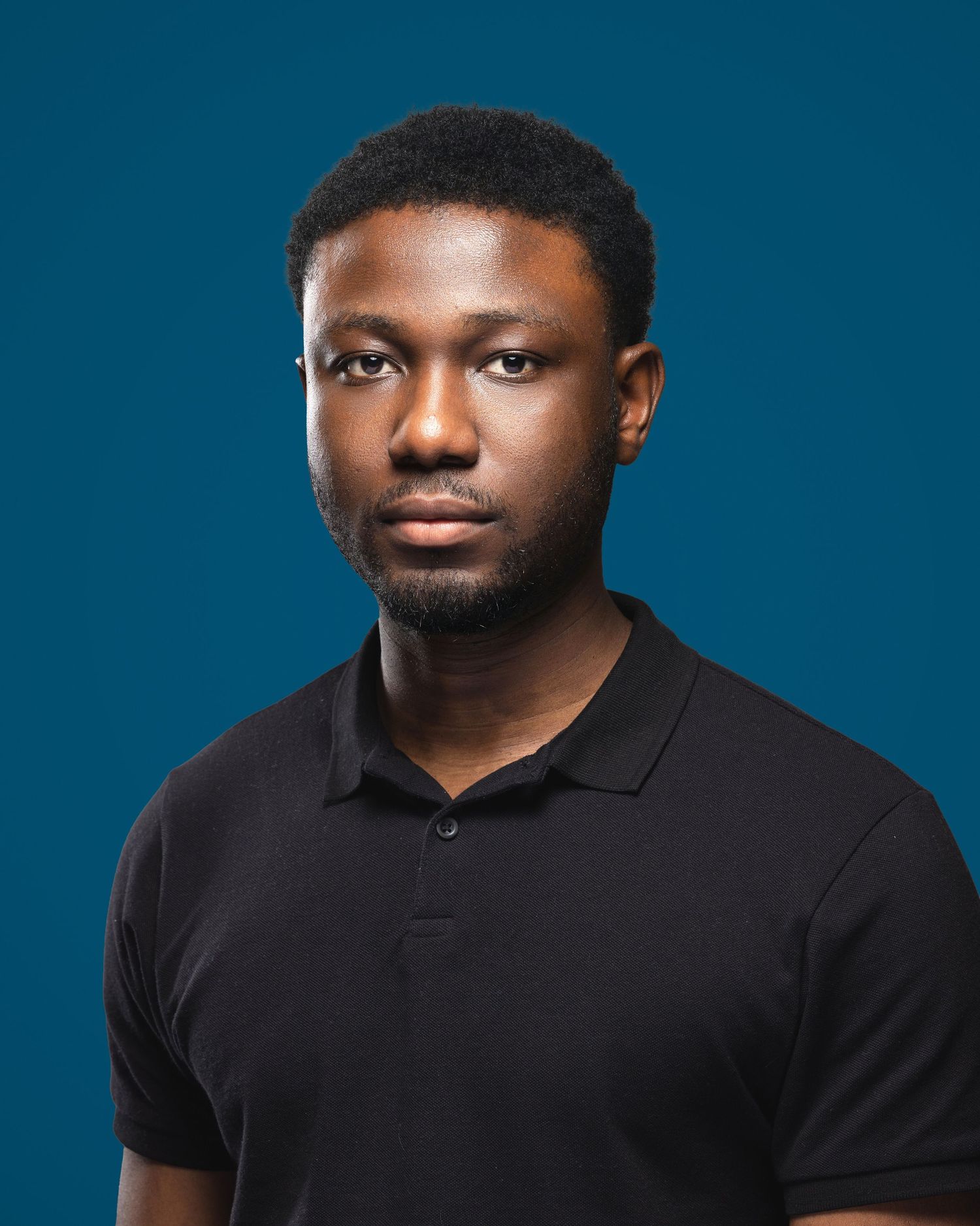Young Black man in black polo shirt poses for professional headshot against teal blue background.