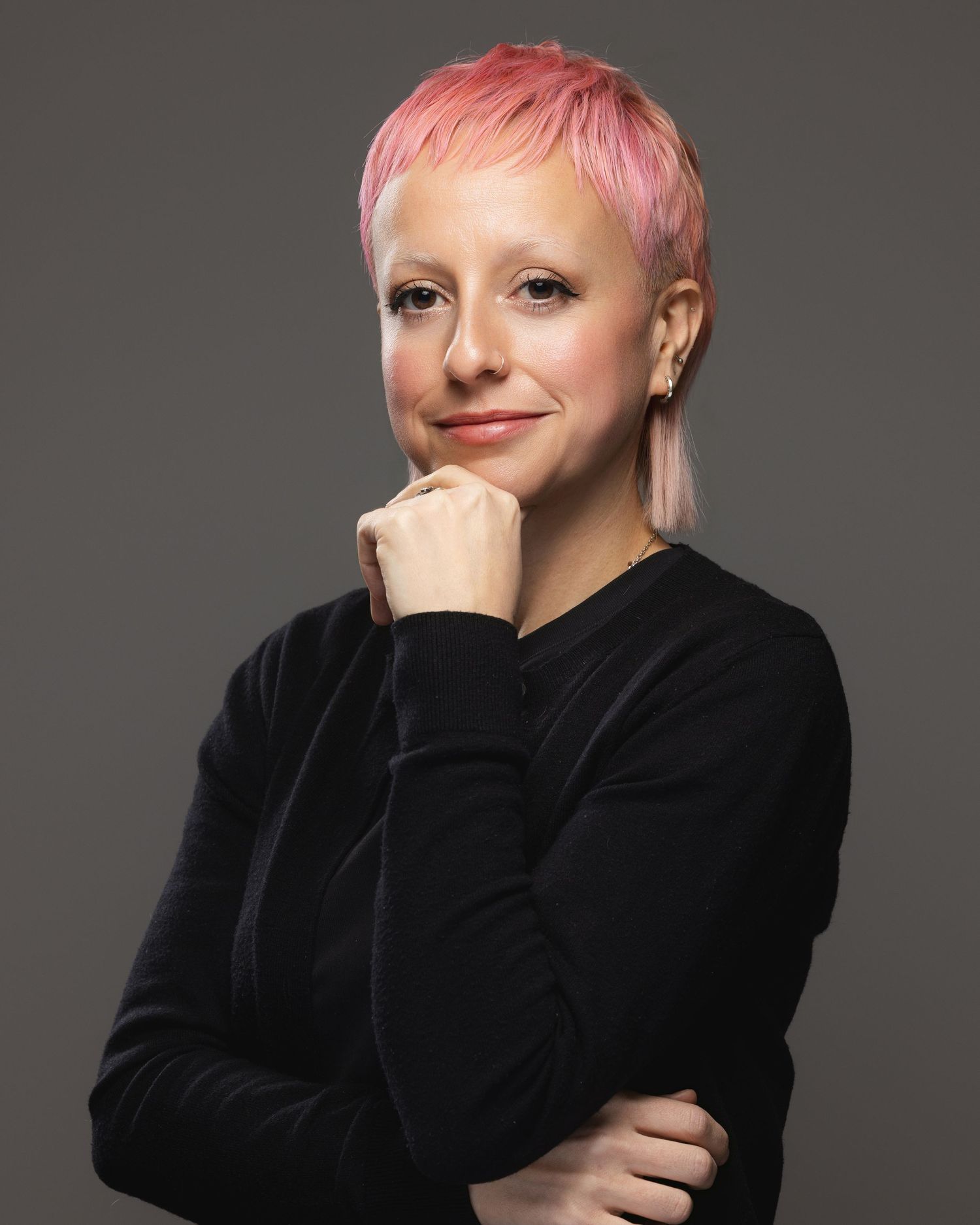 Person with short pink hair wearing black top, posing confidently with hand on chin against gray background.
