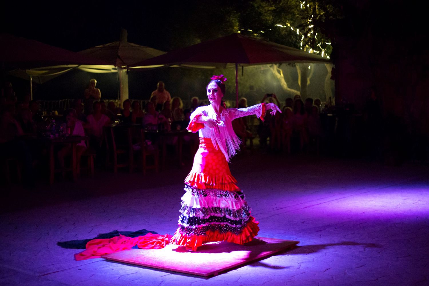 Flamenco dancer in a red and white ruffled dress performs on a wooden platform under purple stage lighting as an outdoor audience watches.