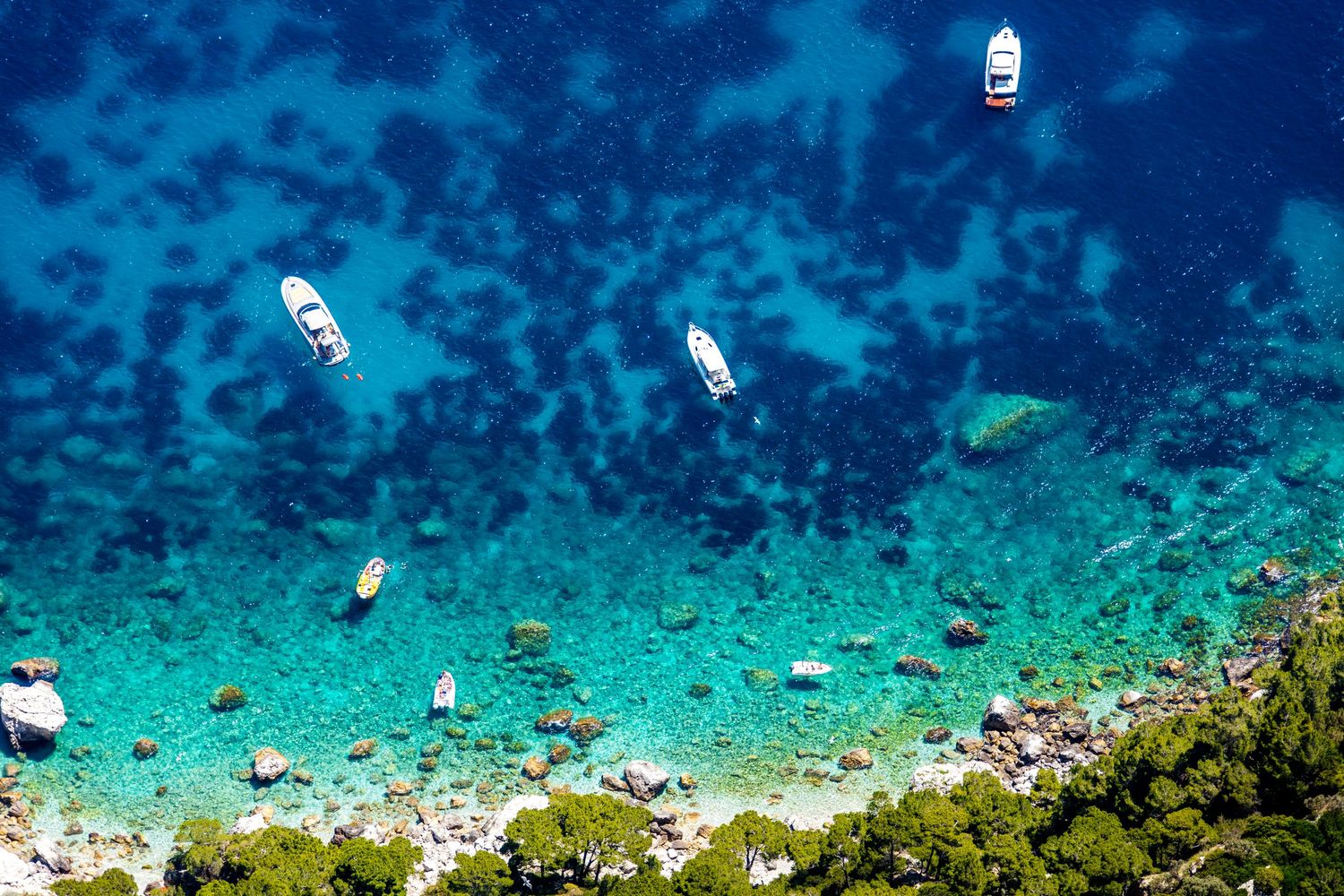 Aerial drone photograph of sailing boats anchored in crystal-clear Mediterranean waters along a rugged rocky coastline.