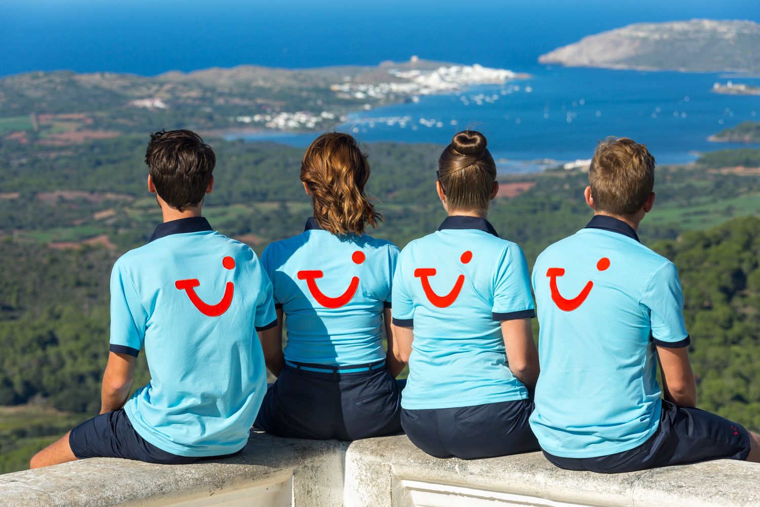 Four people in matching blue TUI shirts with TUI Smile sit on a ledge overlooking a Mediterranean coastal landscape.