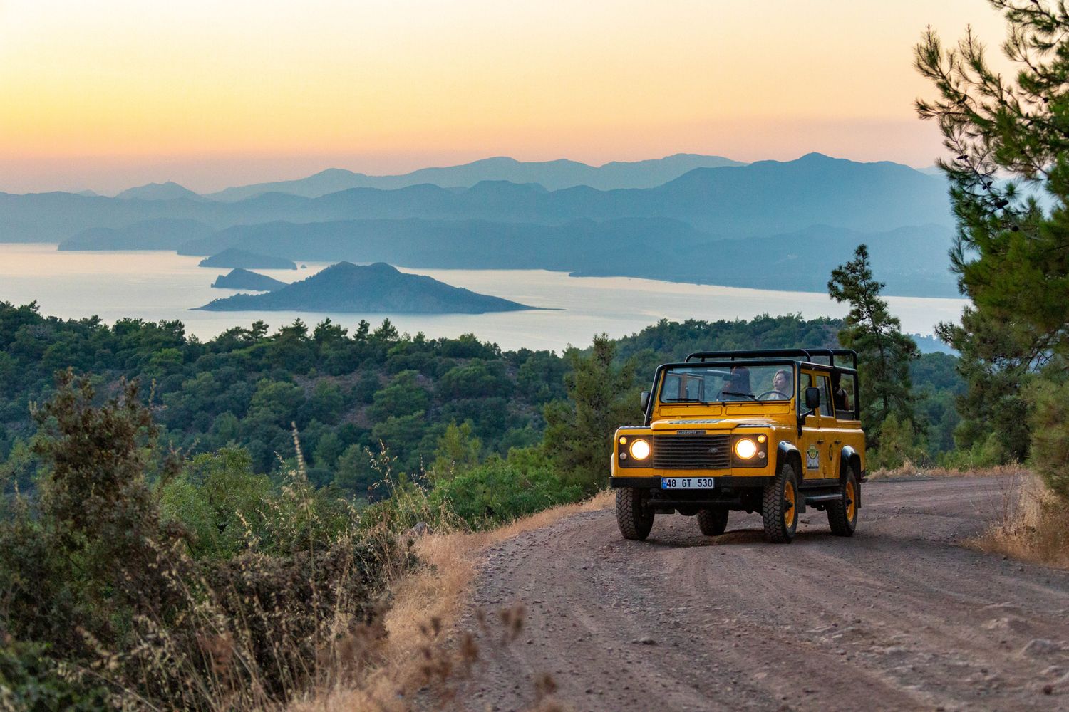 Yellow Land Rover drives on mountain road overlooking scenic coastal islands during golden sunset.