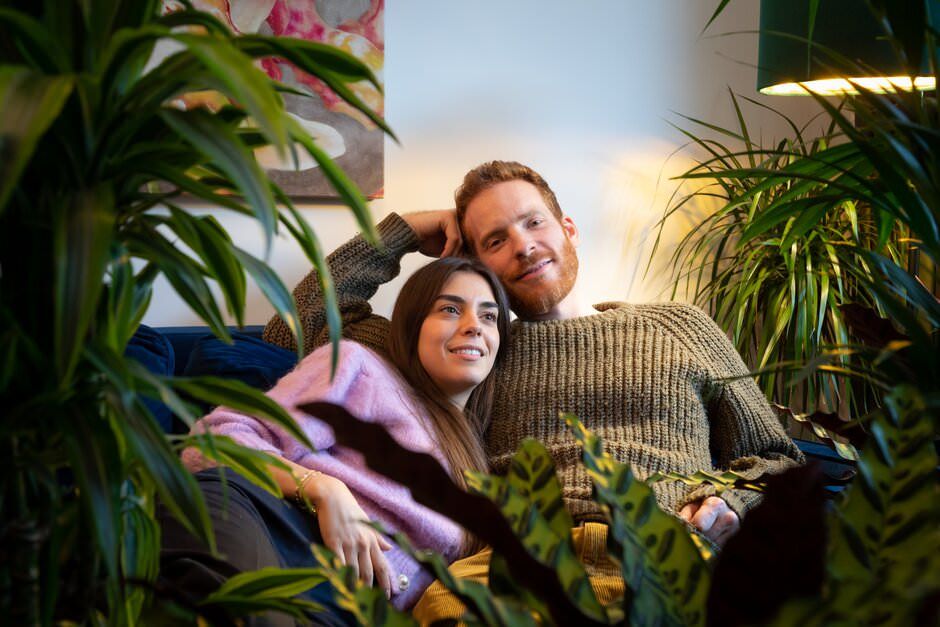 A couple relaxes together surrounded by lush green plants and warm lighting in an indoor garden setting.