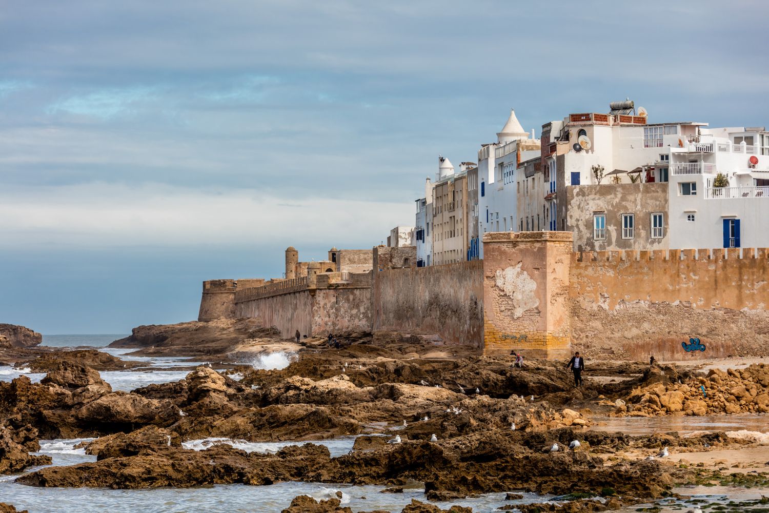 Ancient fortress walls and white buildings of Essaouira medina rise above rocky coastline in Morocco at sunset.
