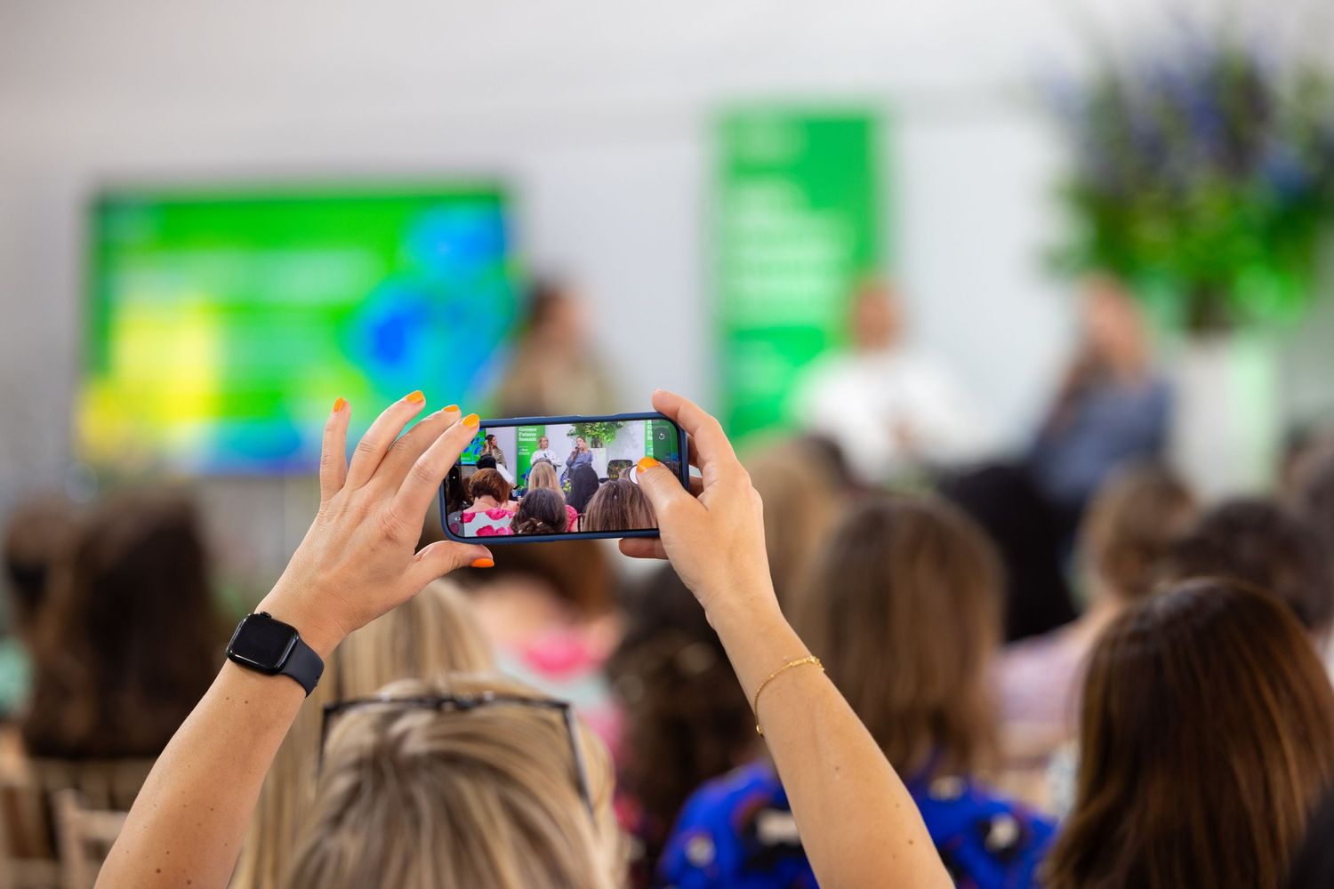 Person holding smartphone to record a panel discussion at a conference with audience seated in rows.