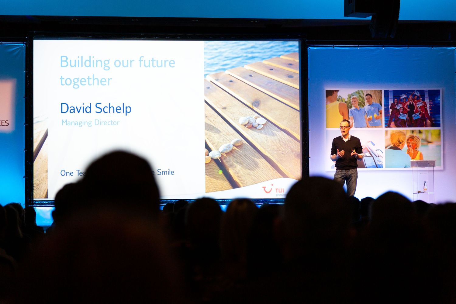 A male speaker presents at a corporate conference podium on a blue-lit stage with artwork displayed in the background.