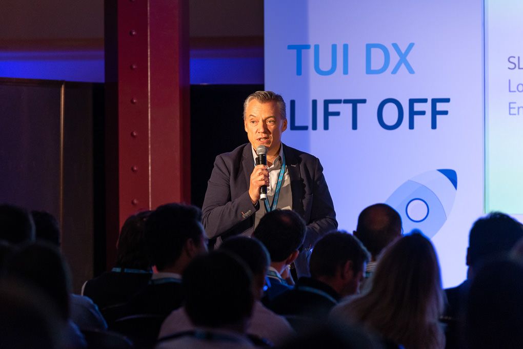 A person speaking at a professional conference with blue lighting and presentation screens visible in the background. Conference photography