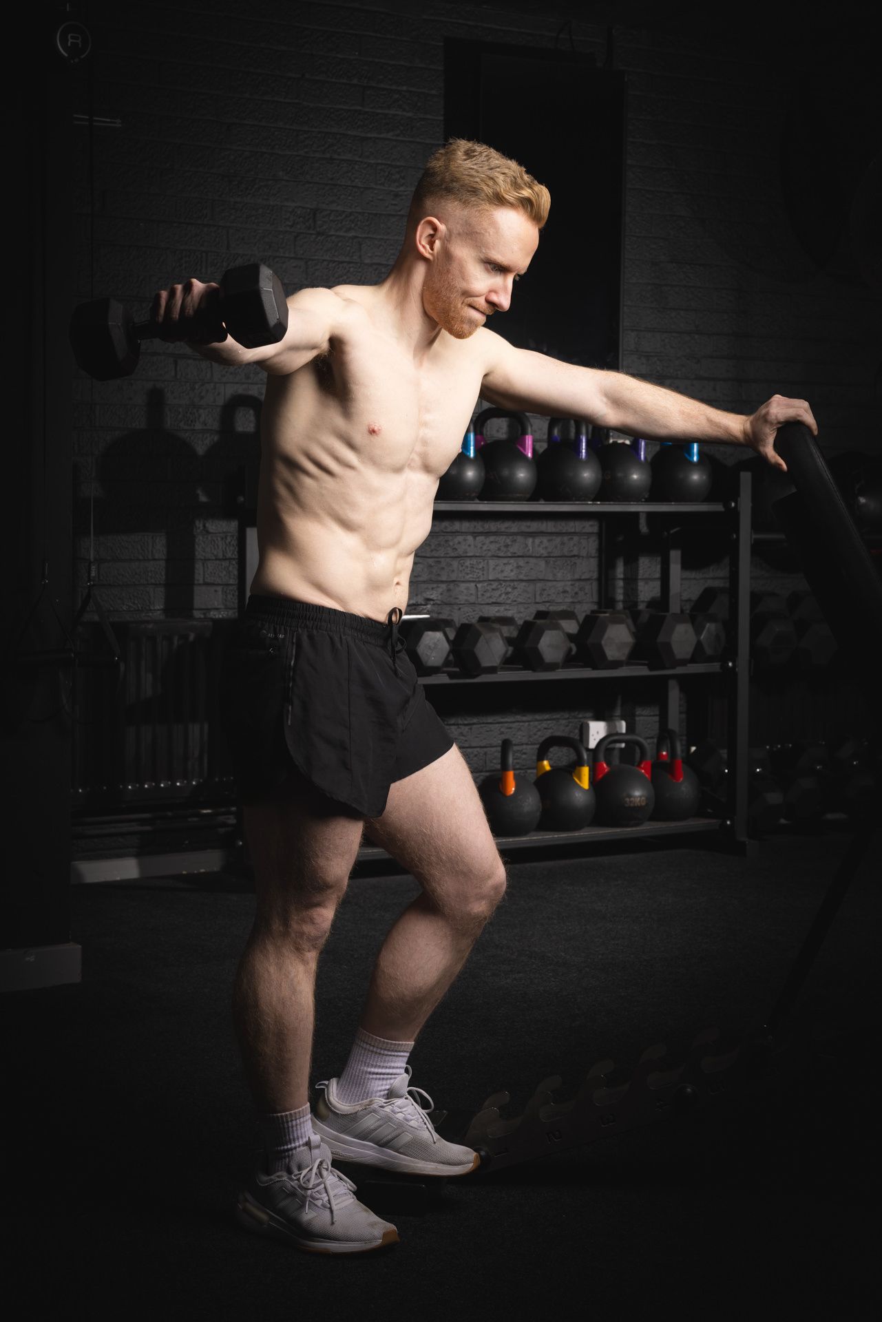 An athletic male performs a dynamic lunging exercise in a dark Hicthin gym setting with kettlebells visible in the background.