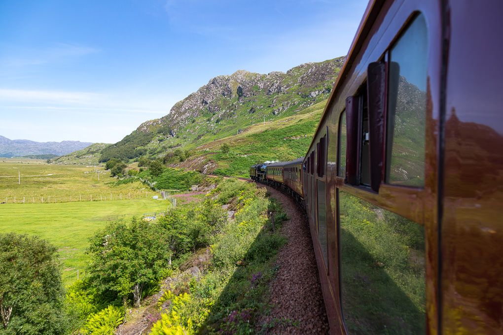 Scenic railway journey on the Jacobite Express through green mountains with vintage train cars winding along hillside tracks.