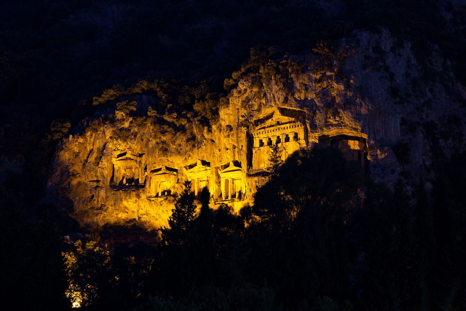 Ancient rock-cut tombs illuminated in golden light against a dark cliffside at night.