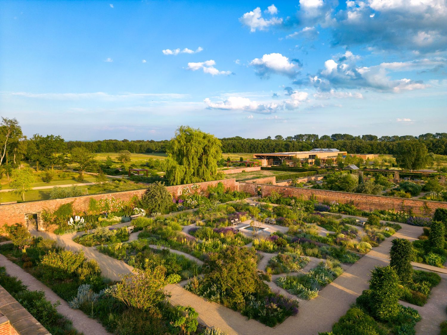 Aerial drone photograph of a formal walled garden featuring geometric pathways, landscaped planting and mature trees under clear blue skies.