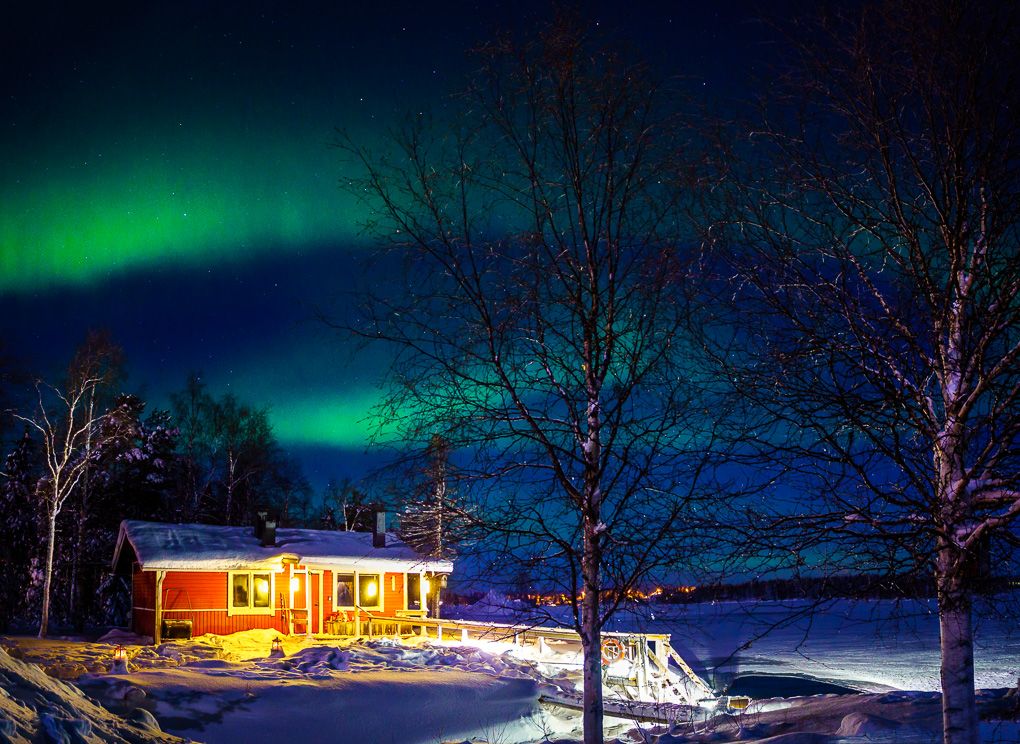Northern lights dance over illuminated cabin in snowy winter landscape with bare trees silhouetted against dark sky.