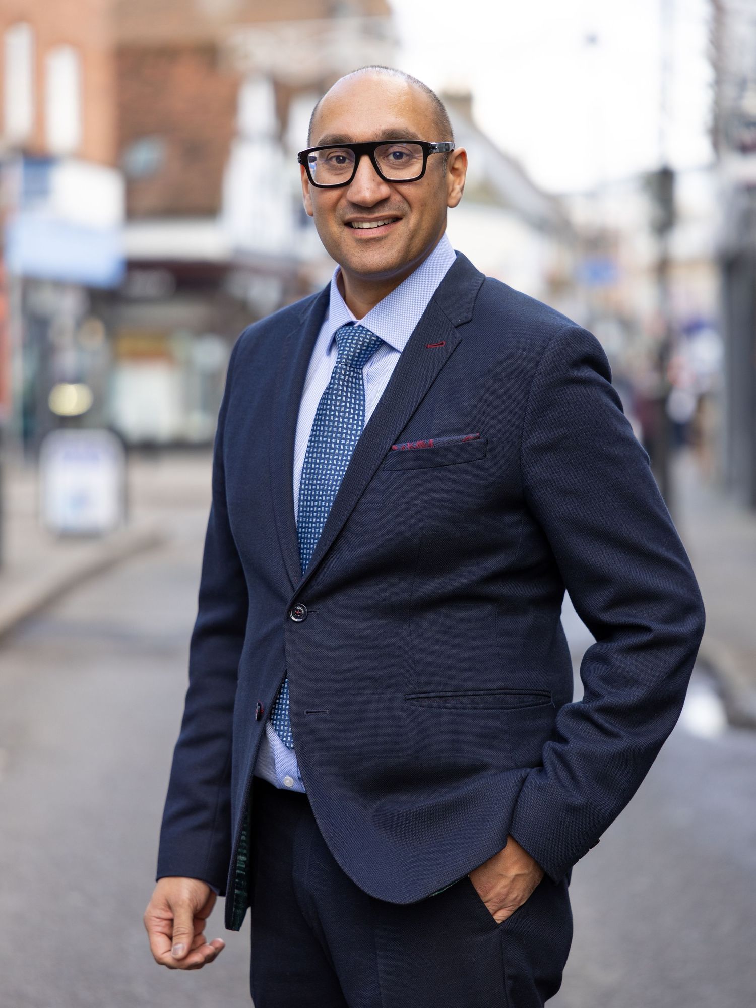 Professional man in navy suit and blue tie standing on a blurred urban street, smiling confidently.