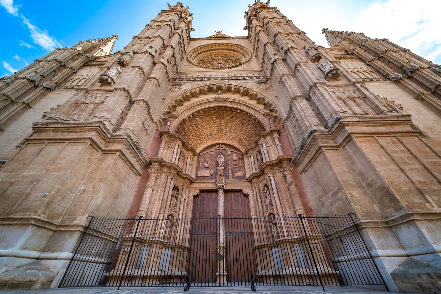 Gothic cathedral facade with ornate stone carvings, large wooden doors, and iron gates under a blue sky.