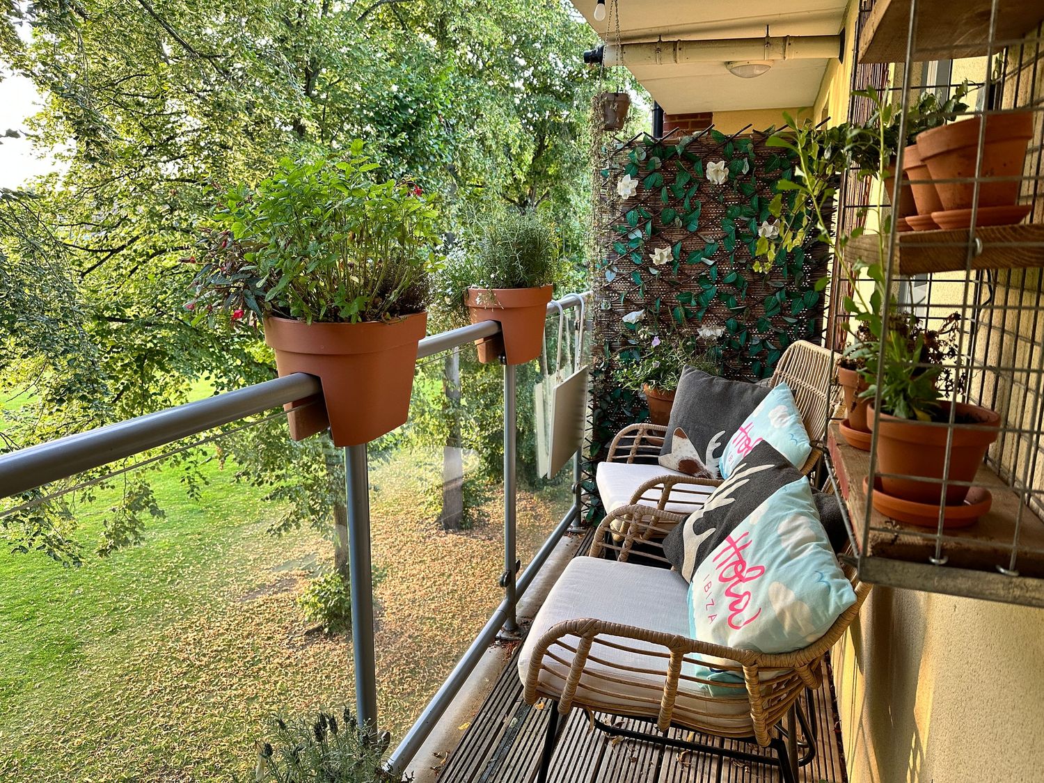 A cozy balcony with potted plants, rattan chairs, colorful cushions, and a floral wall panel overlooking a lush green garden.