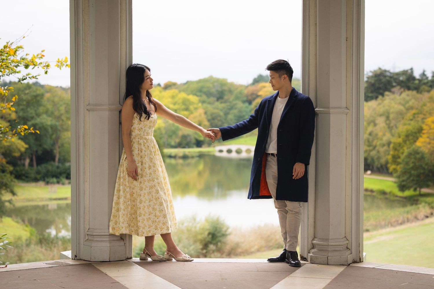 Couple holding hands in elegant outdoor pavilion with scenic lake and autumn trees backdrop.