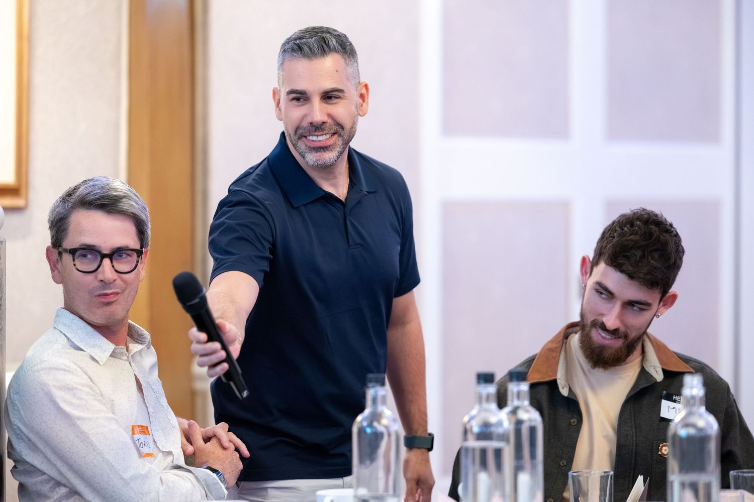 Three men at a conference table, one standing holding a microphone, smiling during a panel discussion.